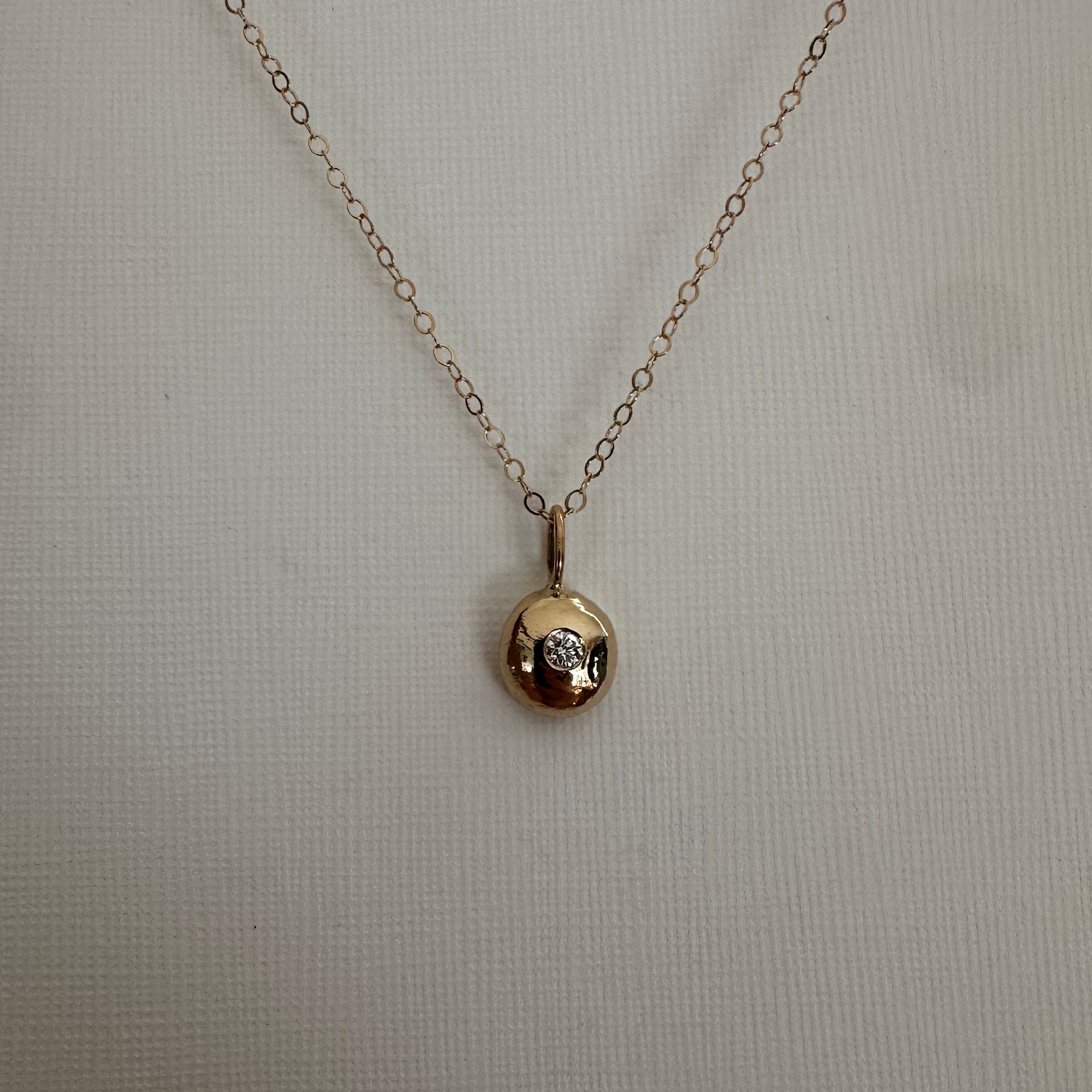 A delicate gold chain necklace with a small, polished gold pendant featuring a tiny clear gemstone, displayed against a textured off-white background.
