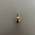 A small gold pendant charm featuring a circular bezel-set dark gemstone, displayed on a textured light gray fabric background.