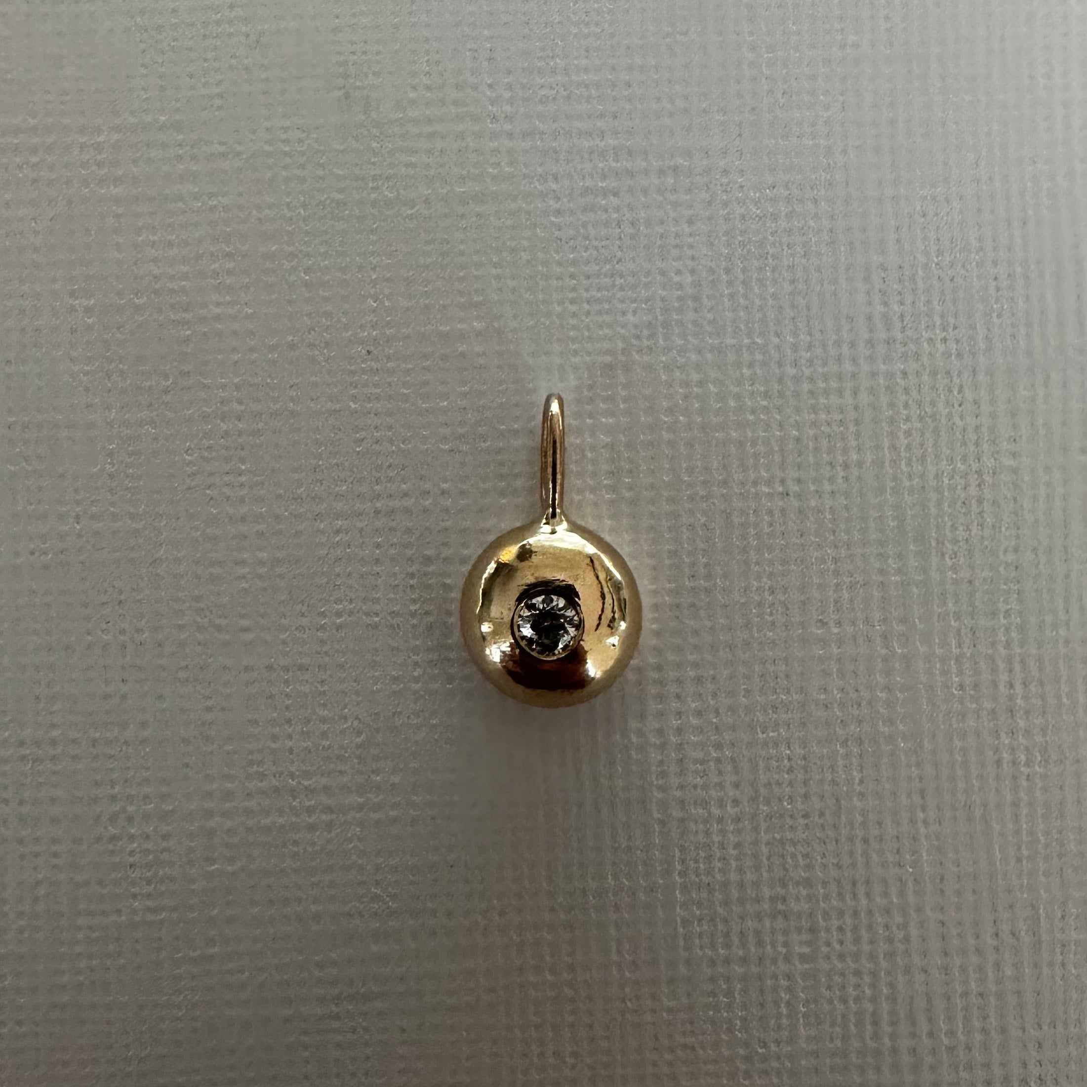 A small gold pendant charm featuring a circular bezel-set dark gemstone, displayed on a textured light gray fabric background.