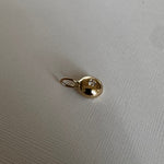 A small gold oval pendant charm with a tiny clear gemstone in the center, resting on a textured off-white fabric surface.