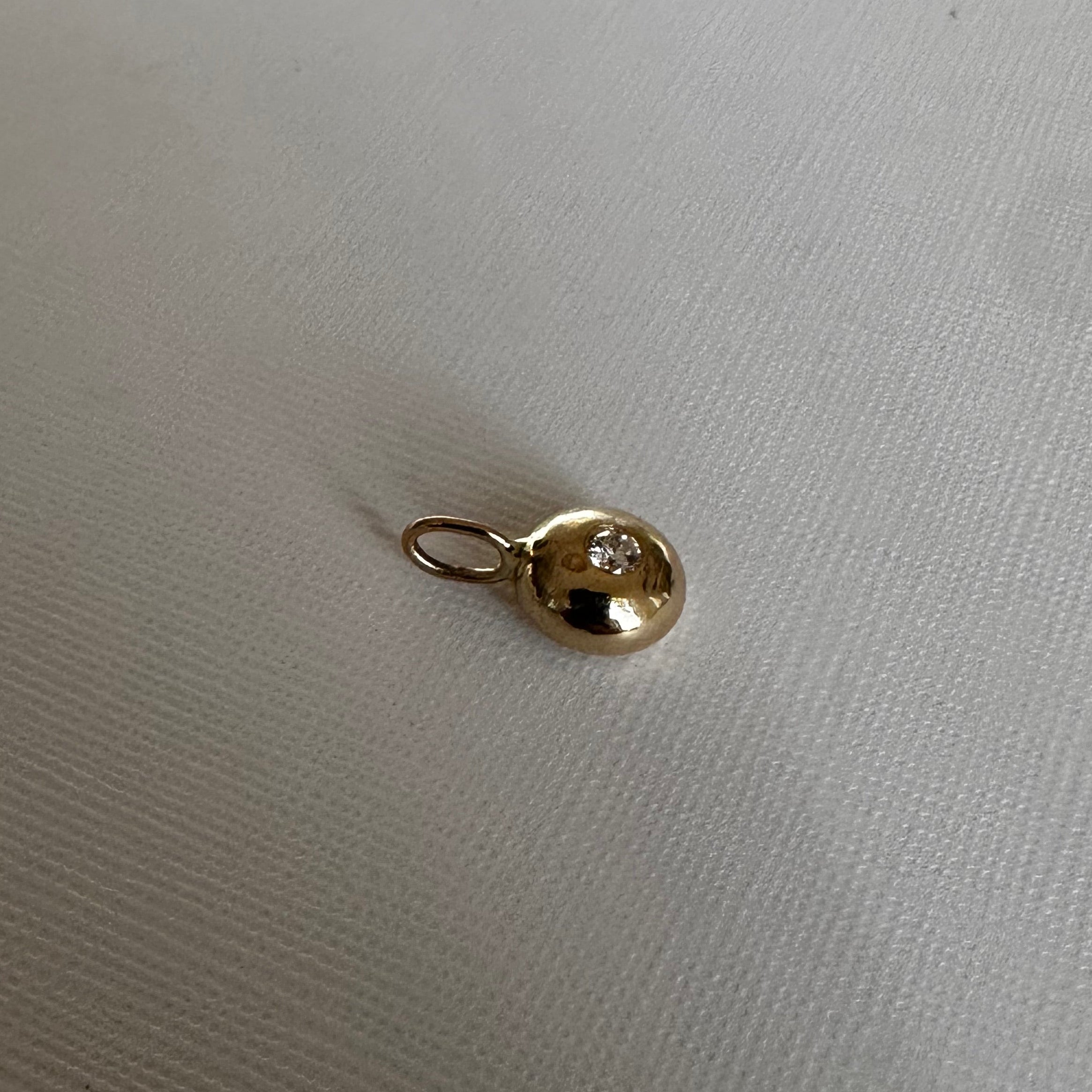 A small gold oval pendant charm with a tiny clear gemstone in the center, resting on a textured off-white fabric surface.