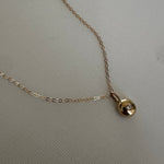A delicate gold chain necklace with a small, polished gold disc pendant featuring a tiny clear gemstone, displayed on a textured light beige fabric background.