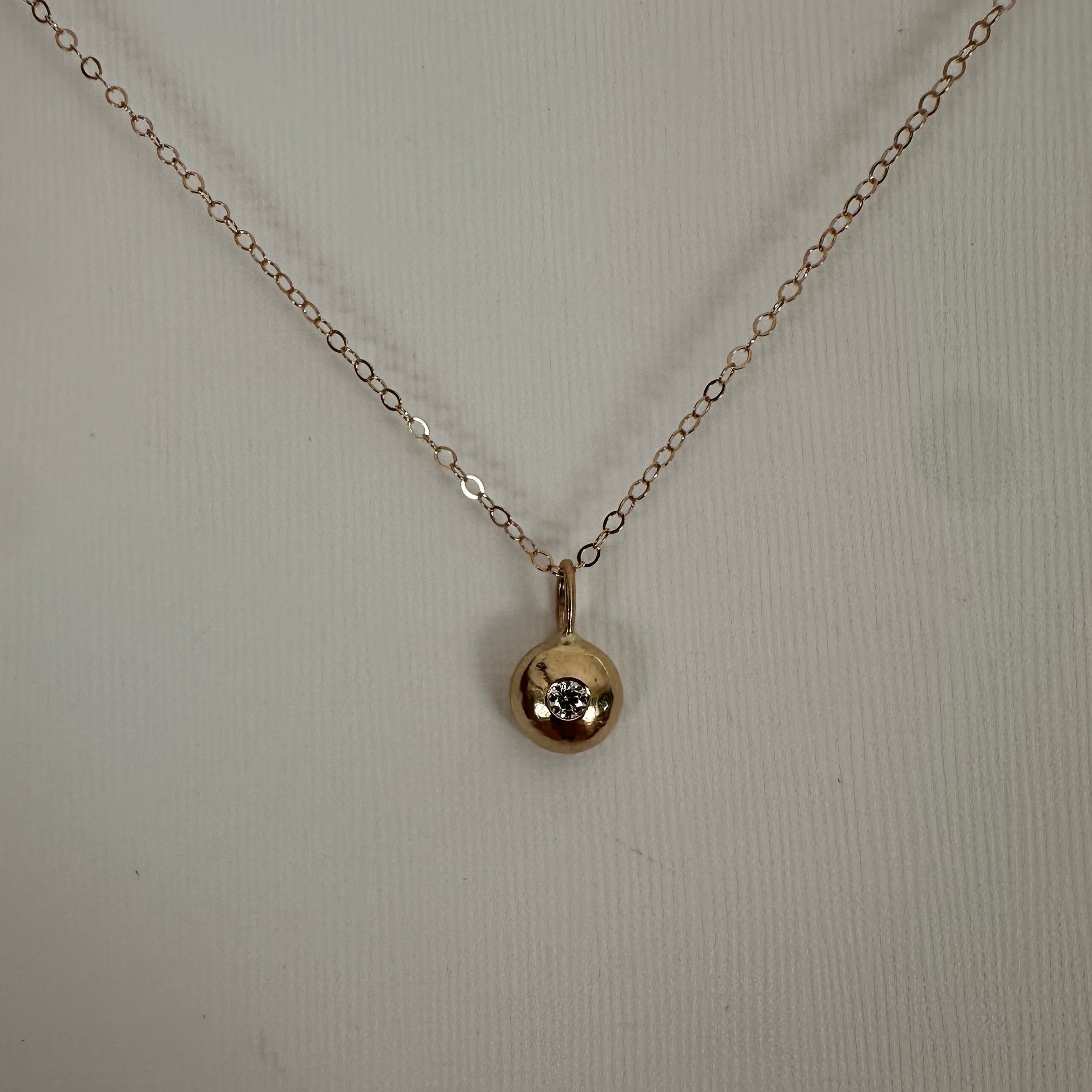 A delicate yellow gold chain necklace with a small spherical pendant containing a tiny clear gemstone, displayed against a light beige textured background.