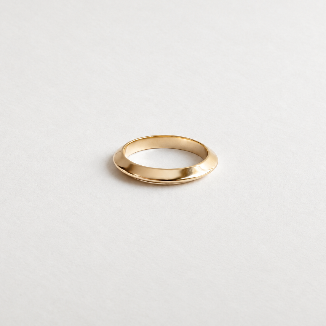 A minimalist gold band ring with a polished, slightly tapered oval shape, photographed on a clean, light gray textured background.