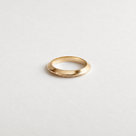 A minimalist gold band ring with a polished, slightly tapered oval shape, photographed on a clean, light gray textured background.