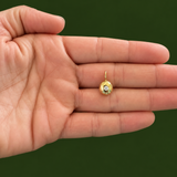 A small gold pendant with a natural diamond is held on the tip of an open hand against a solid dark green background.
