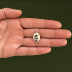 A small oval ring with a silver-toned band and a clear stone featuring a black-and-white portrait, held on the back of an open hand against a dark olive green background.