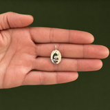 A small oval ring with a silver-toned band and a clear stone featuring a black-and-white portrait, held on the back of an open hand against a dark olive green background.