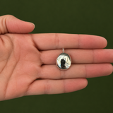 A small oval silver pendant with a reflective metallic surface, held in the center of an open hand against a dark green background.