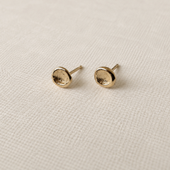 A pair of small gold stud earrings with a textured, hammered circular face, resting on a textured beige linen fabric background.