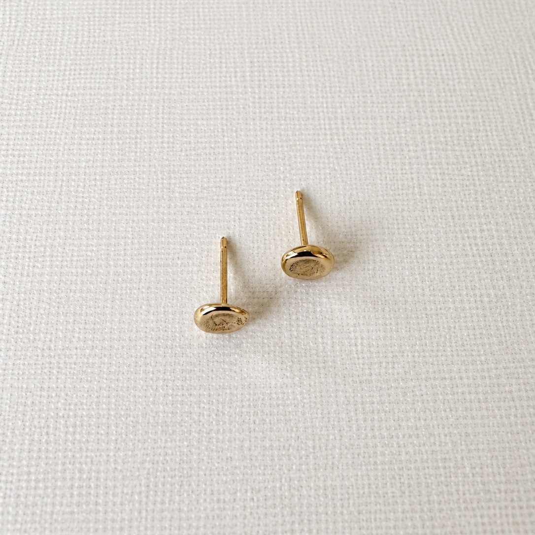 A pair of small gold stud earrings with oval disc fronts and straight posts, displayed on a textured off-white fabric background.
