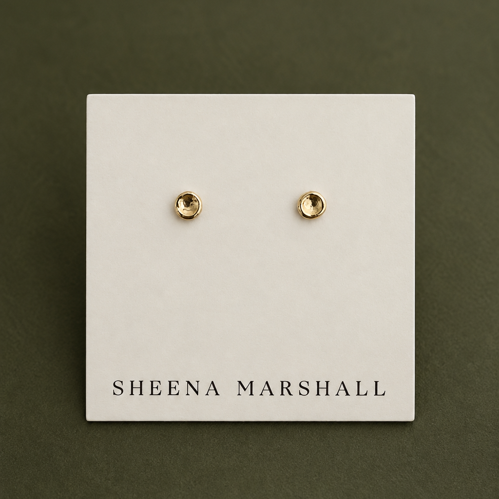 A pair of small gold stud earrings displayed on a white card labeled SHEENA MARSHALL, set against a dark olive green background.
