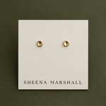 A pair of small gold stud earrings displayed on a white card labeled SHEENA MARSHALL, set against a dark olive green background.