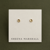 A pair of small gold stud earrings displayed on a white card labeled SHEENA MARSHALL, set against a dark olive green background.
