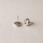 A pair of silver stud earrings with flat circular faces, shown from the back on a textured off-white fabric background.