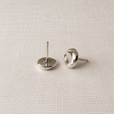 A pair of silver stud earrings with flat circular faces, shown from the back on a textured off-white fabric background.