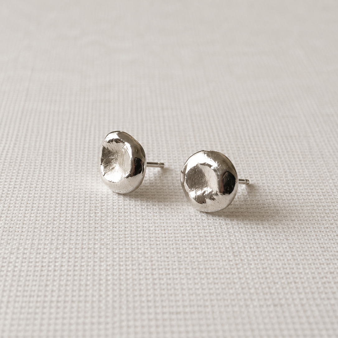 A pair of silver stud earrings with a textured, organic disc-like shape, displayed on a light beige woven fabric background.