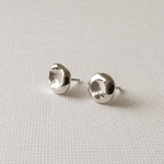 A pair of silver stud earrings with a textured, organic disc-like shape, displayed on a light beige woven fabric background.