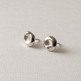A pair of silver stud earrings with a textured, organic disc-like shape, displayed on a light beige woven fabric background.
