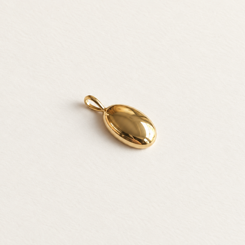 A small polished gold oval pendant with a loop at the top, photographed on a clean off-white textured background.