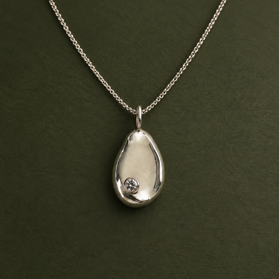 A silver teardrop pendant necklace with a small clear gemstone set in the lower left, displayed on a dark olive green background.