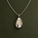 A silver teardrop pendant necklace with a small clear gemstone set in the lower left, displayed on a dark olive green background.