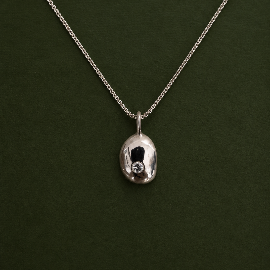A silver chain necklace with a polished, organic-shaped silver pendant featuring a small clear gemstone, displayed against a dark green background.