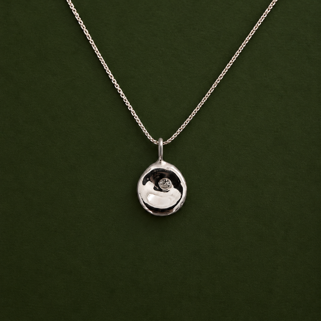 A silver pendant necklace with a small, polished circular disc pendant on a delicate chain, displayed against a dark green background.