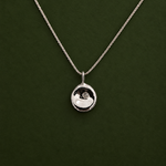 A silver pendant necklace with a small, polished circular disc pendant on a delicate chain, displayed against a dark green background.
