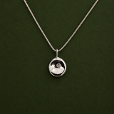 A silver pendant necklace with a small, polished circular disc pendant on a delicate chain, displayed against a dark green background.