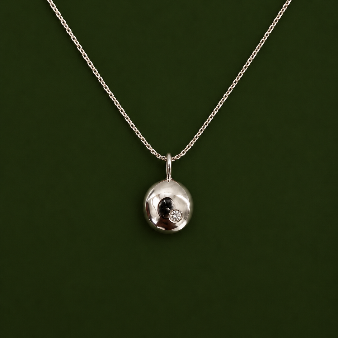 A silver chain necklace with a small, polished silver oval pendant featuring a black and white yin-yang symbol and a tiny clear gemstone, set against a dark green background.