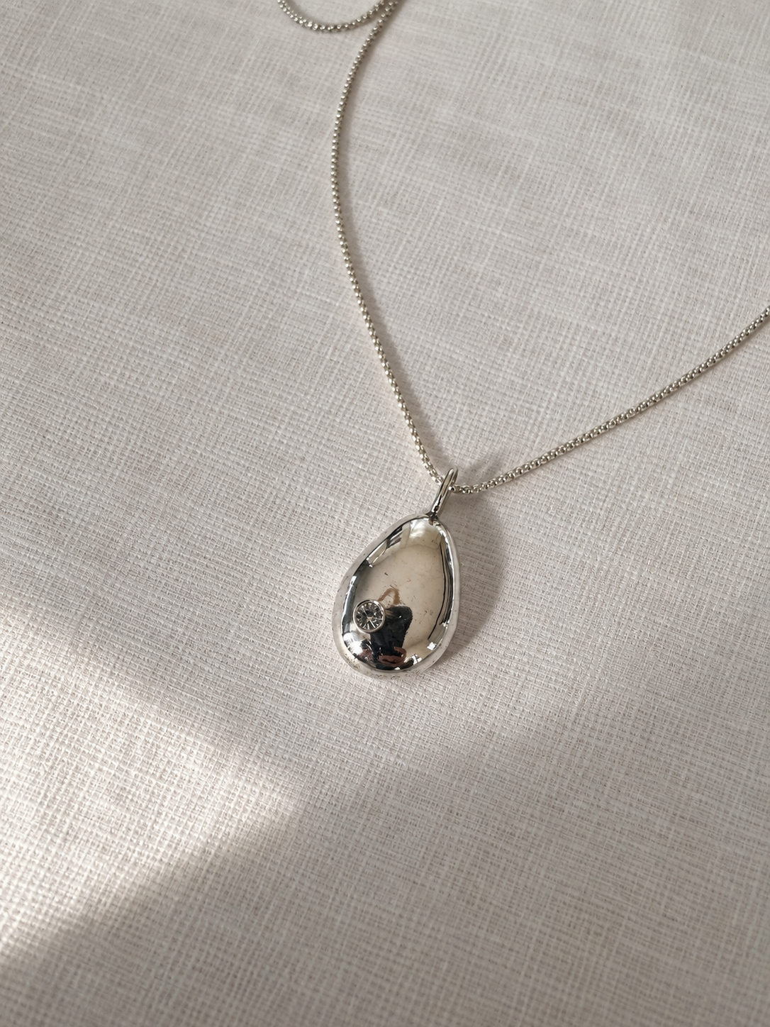 A silver teardrop pendant necklace with a polished, reflective finish, resting on a textured beige linen fabric background.