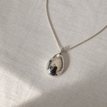 A silver teardrop pendant necklace with a polished, reflective finish, resting on a textured beige linen fabric background.
