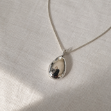 A silver teardrop pendant necklace with a polished, reflective finish, resting on a textured beige linen fabric background.