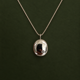 A silver oval pendant necklace with a small clear gemstone, shown on a dark green textured background.