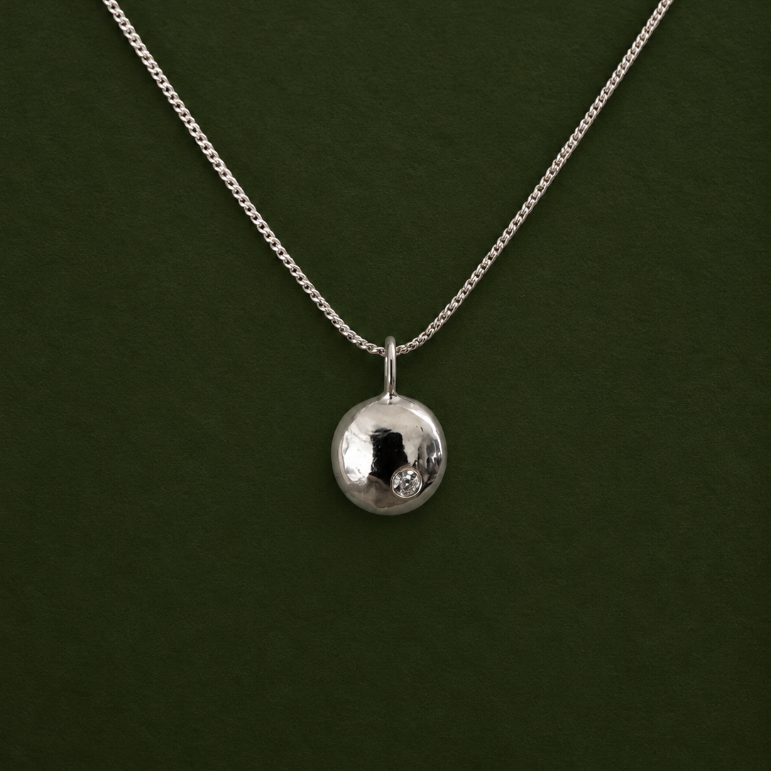 A silver chain necklace with a small, polished silver disc pendant, displayed against a solid dark green background.