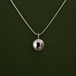 A silver chain necklace with a small, polished silver disc pendant, displayed against a solid dark green background.