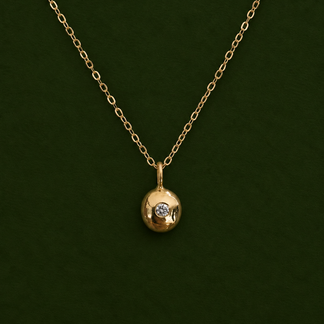 A gold chain necklace with a small round gold pendant featuring a single clear gemstone, displayed against a dark green background.