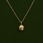 A gold chain necklace with a small round gold pendant featuring a single clear gemstone, displayed against a dark green background.