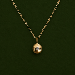 A gold chain necklace with a small round gold pendant featuring a single clear gemstone, displayed against a dark green background.