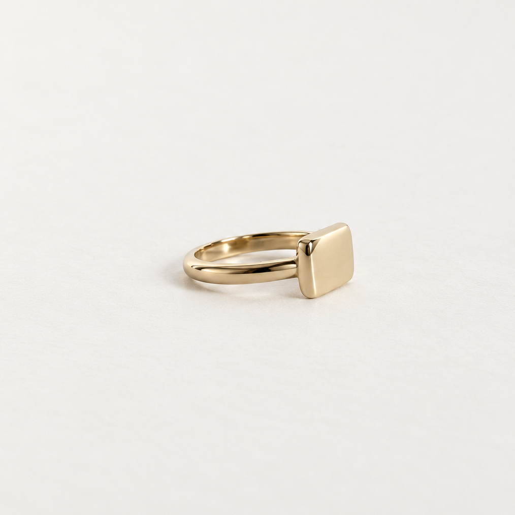 A minimalist gold signet ring with a flat, polished rectangular face, displayed on a clean, light off-white background.