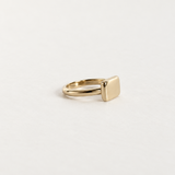 A minimalist gold signet ring with a flat, polished rectangular face, displayed on a clean, light off-white background.