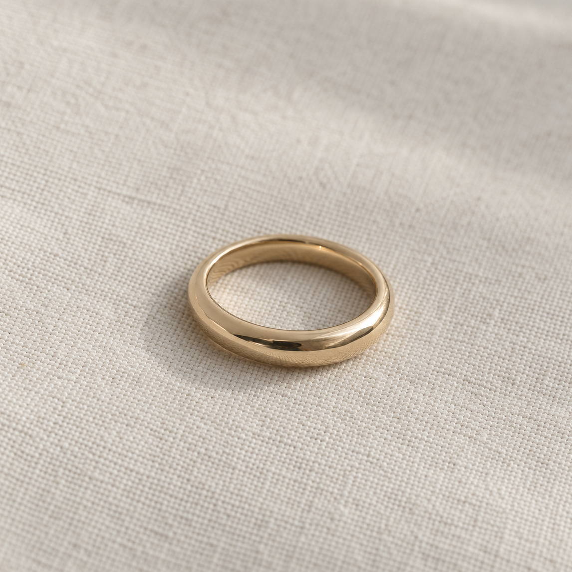 A simple polished gold band ring resting on a textured beige linen fabric background.