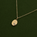 A gold oval pendant necklace with a small clear gemstone set in the lower left, displayed on a dark green textured background.