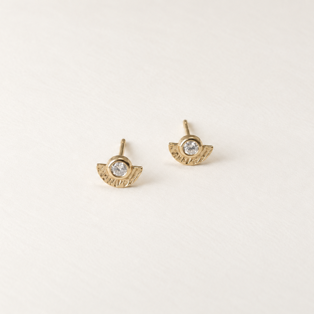 A pair of gold wing stud earrings with a clear round gemstone in the center, displayed on a clean, light off-white background.