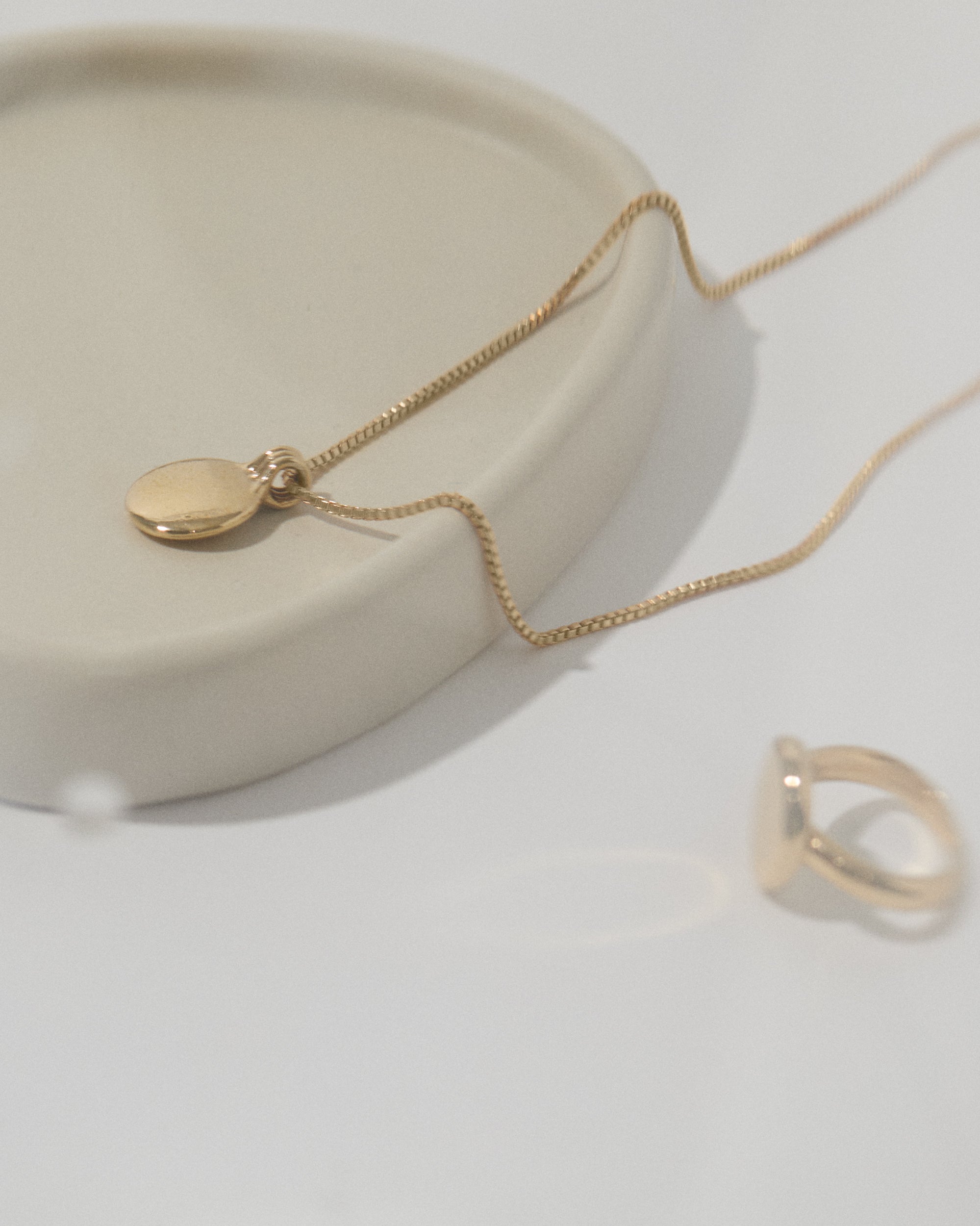 Sustainable fairmined gold jewelry collection featuring delicate disc pendant necklace and minimalist ring, styled on white ceramic surface for ethical fine jewelry showcase