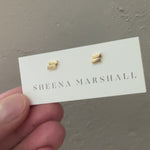 Close-up of solid gold Off-Balance Bar Stud Earrings with an asymmetrical design, symbolizing resilience and balance during life's uncertain moments, displayed on a Sheena Marshall jewelry card.