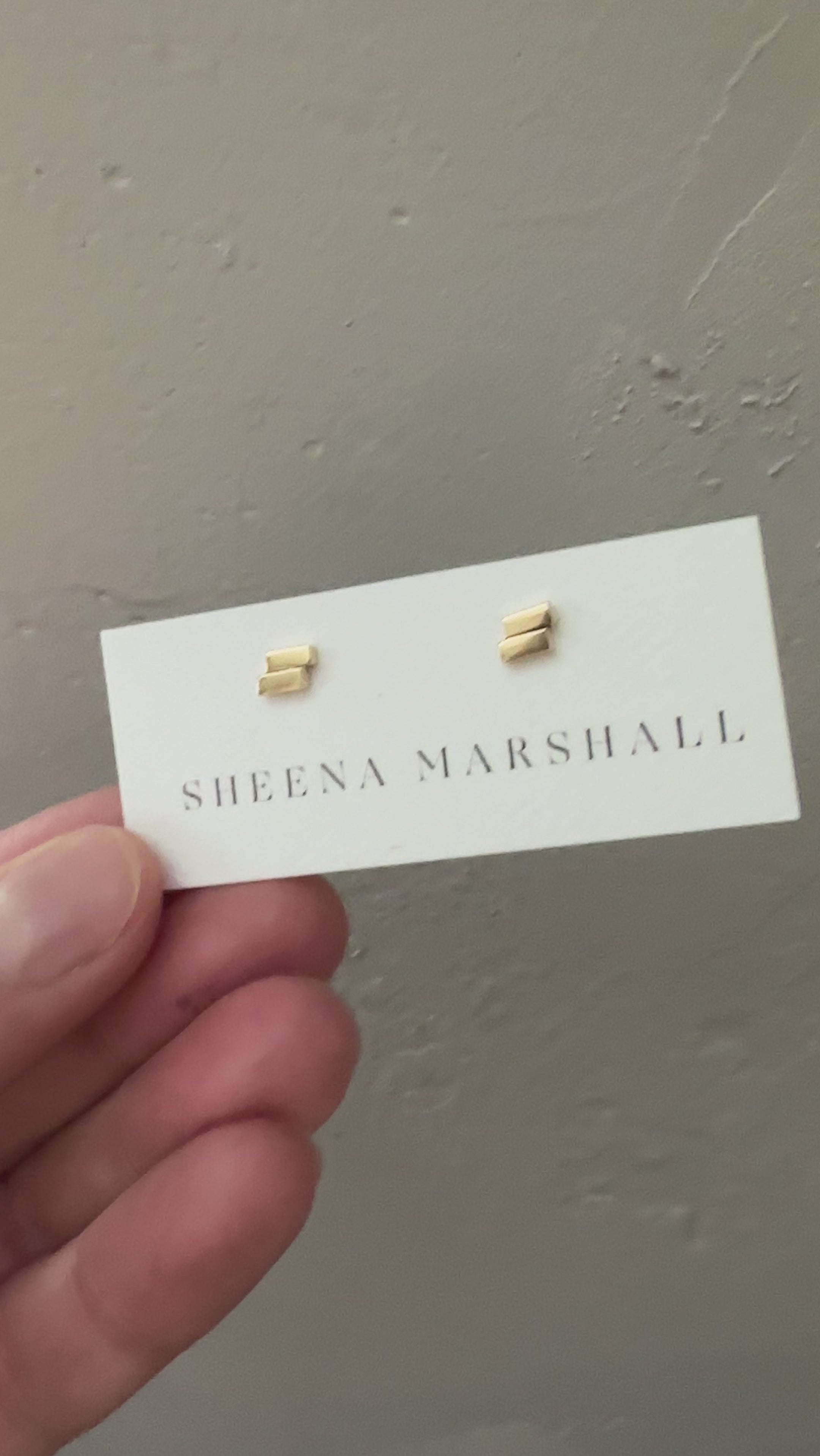 Close-up of solid gold Off-Balance Bar Stud Earrings with an asymmetrical design, symbolizing resilience and balance during life's uncertain moments, displayed on a Sheena Marshall jewelry card.