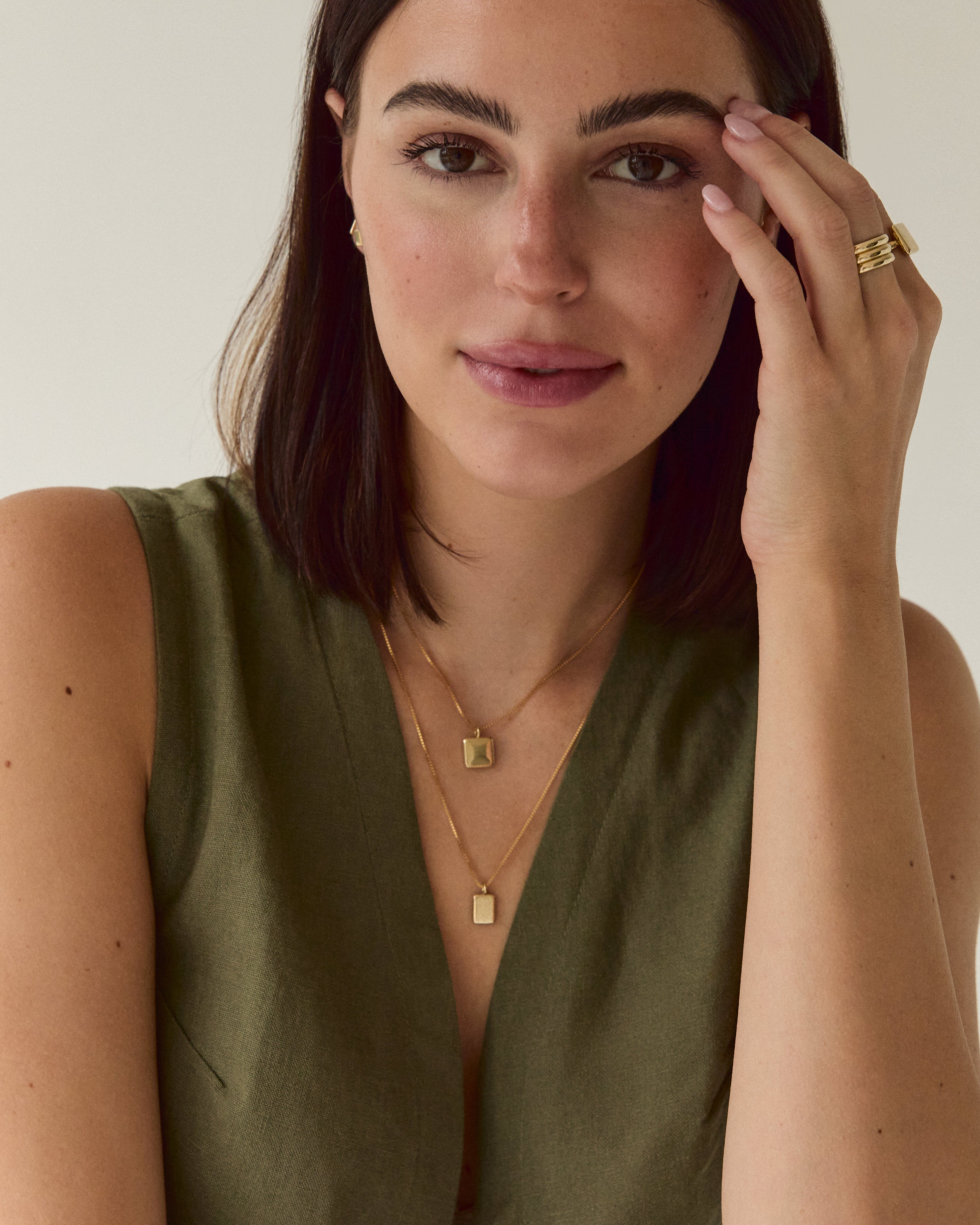 A close-up of a woman wearing a sleeveless olive green top and layered gold necklaces, with gold rings on her fingers, set against a plain light background.